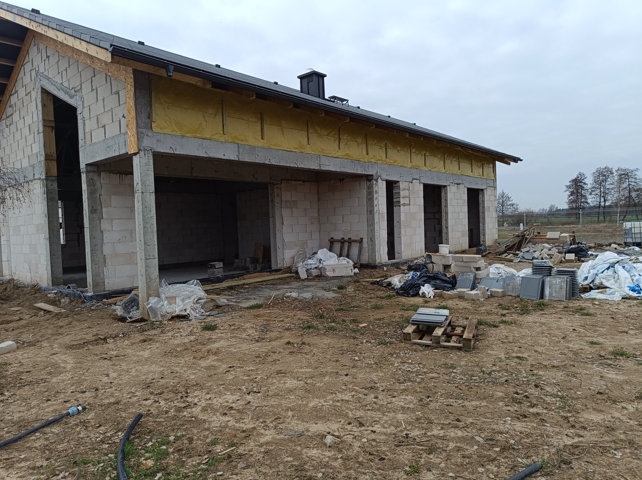 Surowy stan budynku jednorodzinnego z betonu komórkowego z widoczną izolacją stropu i przygotowanymi płytkami elewacyjnymi na palecie, na nierównym, gliniastym terenie budowy.