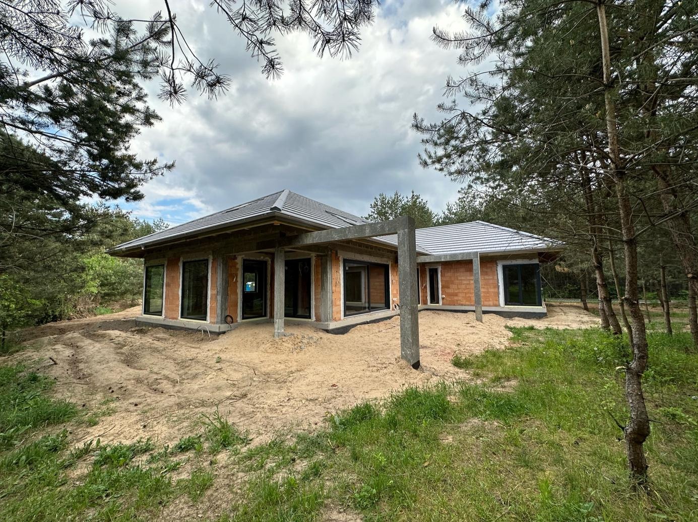 Nowoczesny dom w stanie surowym otwartym, z widoczną konstrukcją z betonu i cegły, otoczony piaszczystym terenem i młodym lasem sosnowym w pochmurny dzień.