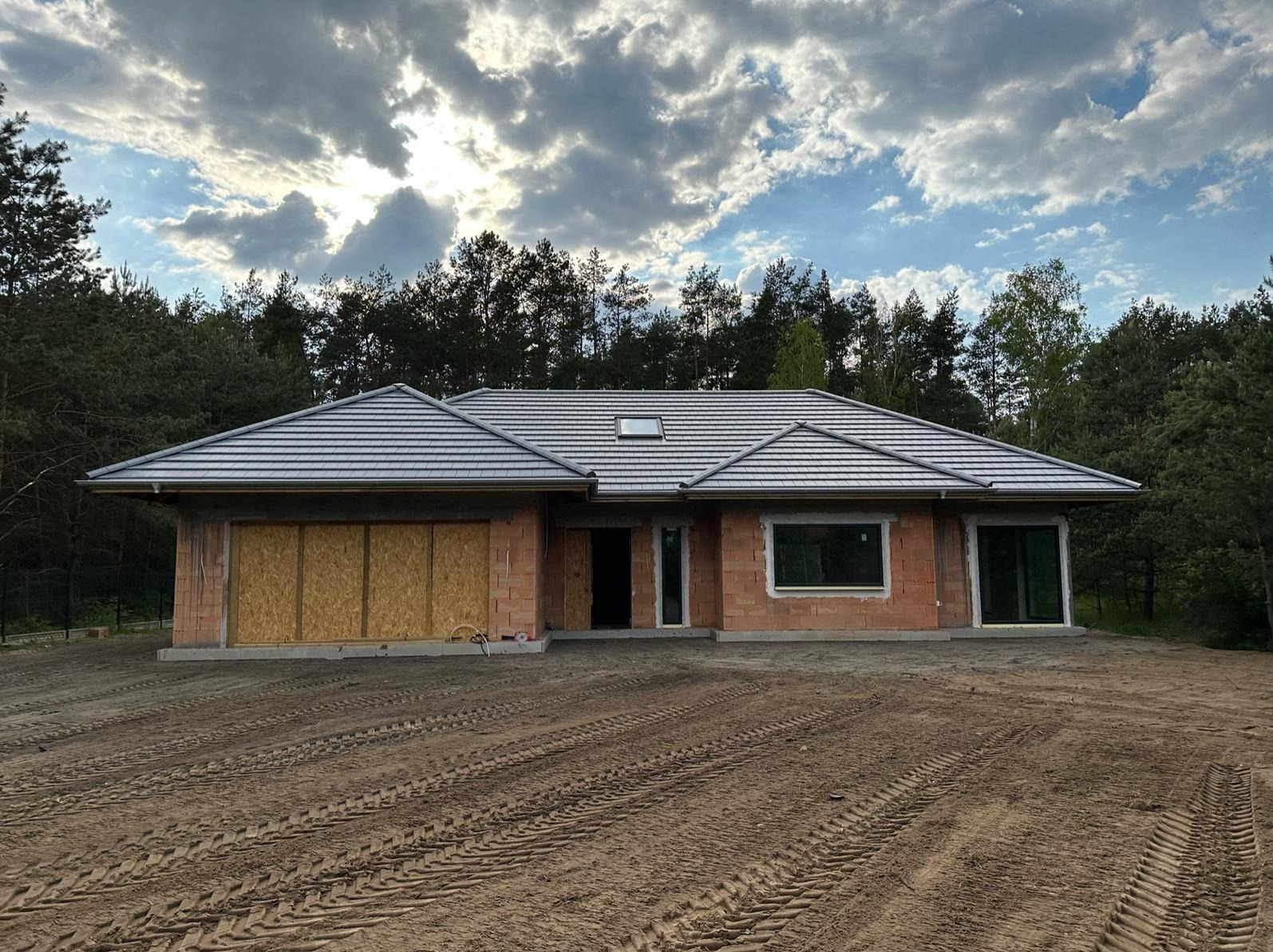 Dom jednorodzinny w trakcie budowy, z widoczną surową elewacją z cegły, szarą dachówką i zamurowanymi otworami garażowymi, na tle lasu i zachmurzonego nieba.
