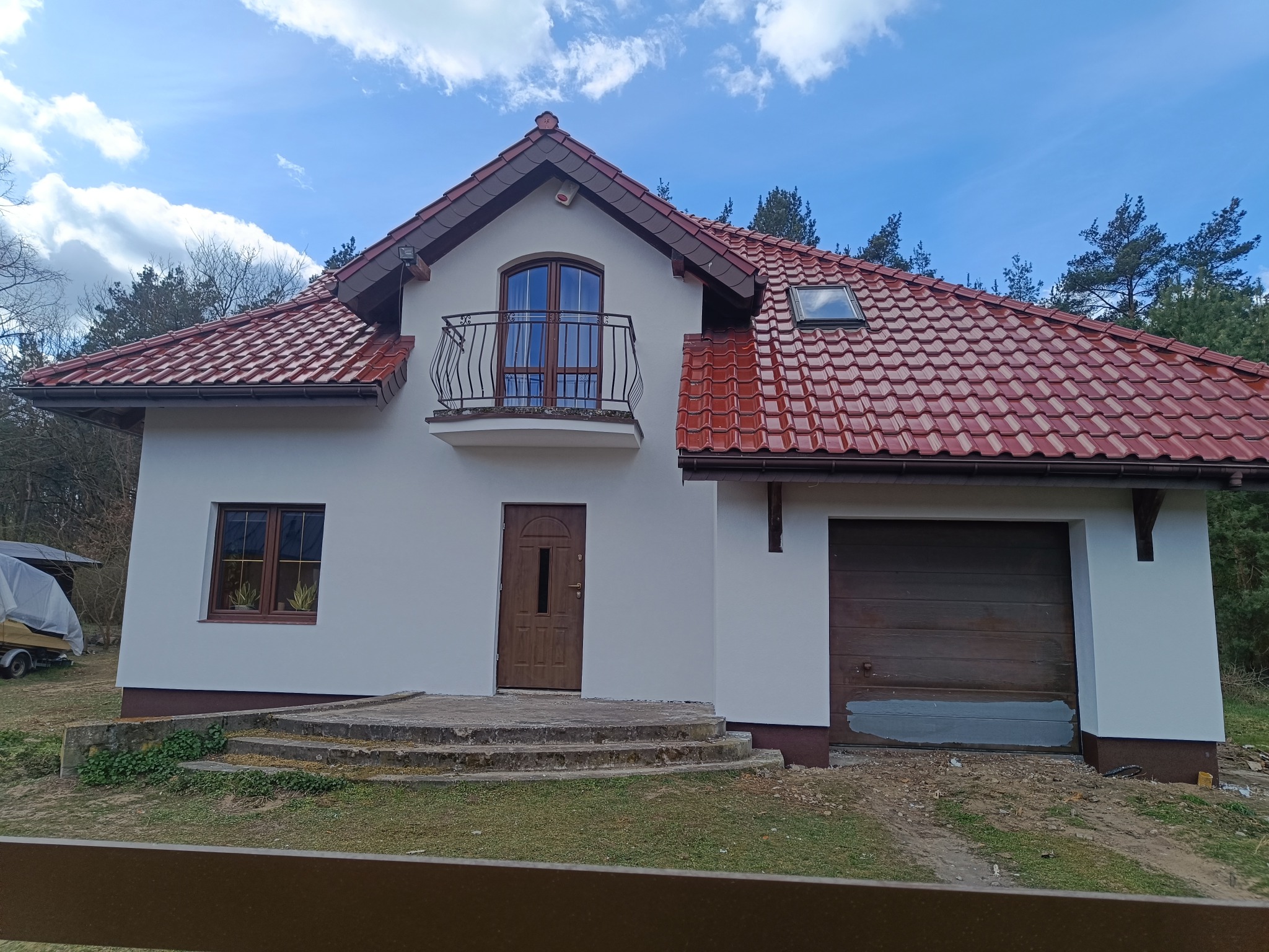 Biały dom jednorodzinny z brązowymi oknami, drzwiami i czerwoną dachówką, widziany z poziomu ogrodzenia. Na piętrze balkon z kutą balustradą. Błękitne niebo w tle.