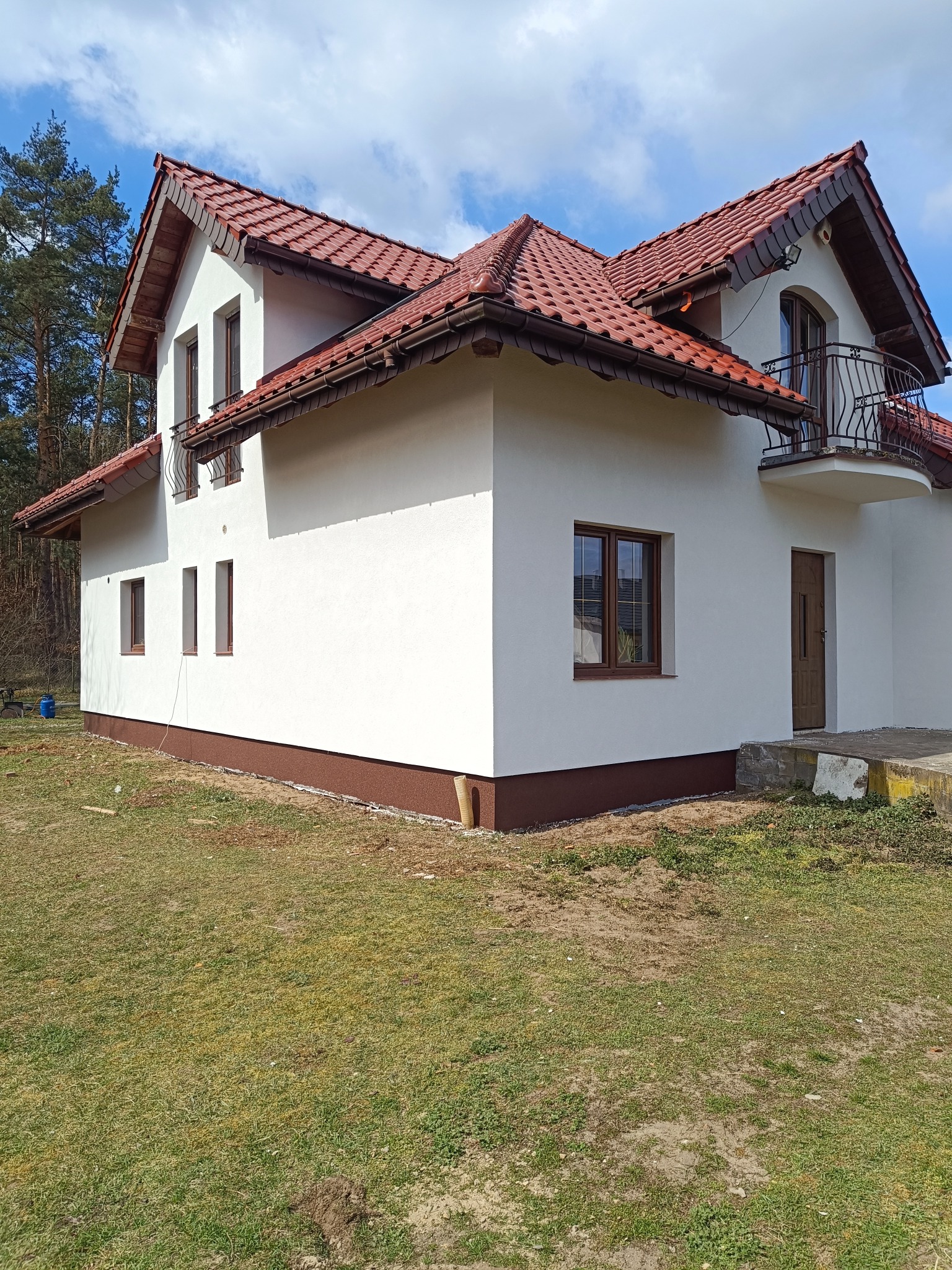 Biały dom z czerwoną dachówką i brązowymi oknami, świeżo po wykonaniu elewacji, widok z boku na tle drzew i błękitnego nieba z chmurami.