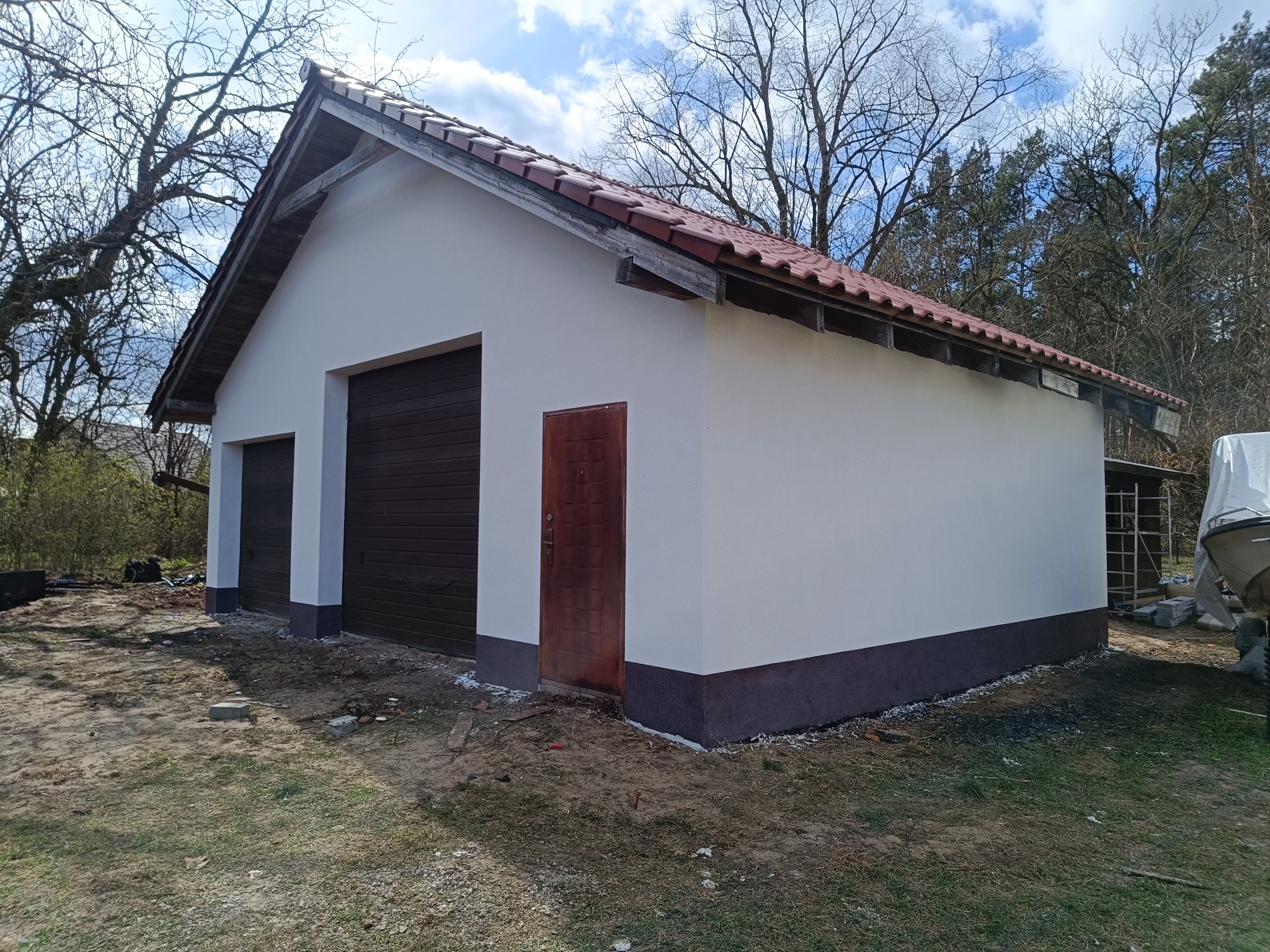 Biały budynek gospodarczy z brązowymi drzwiami garażowymi i wejściowymi, wykończony czerwoną dachówką, na tle drzew bezlistnych i błękitnego nieba.