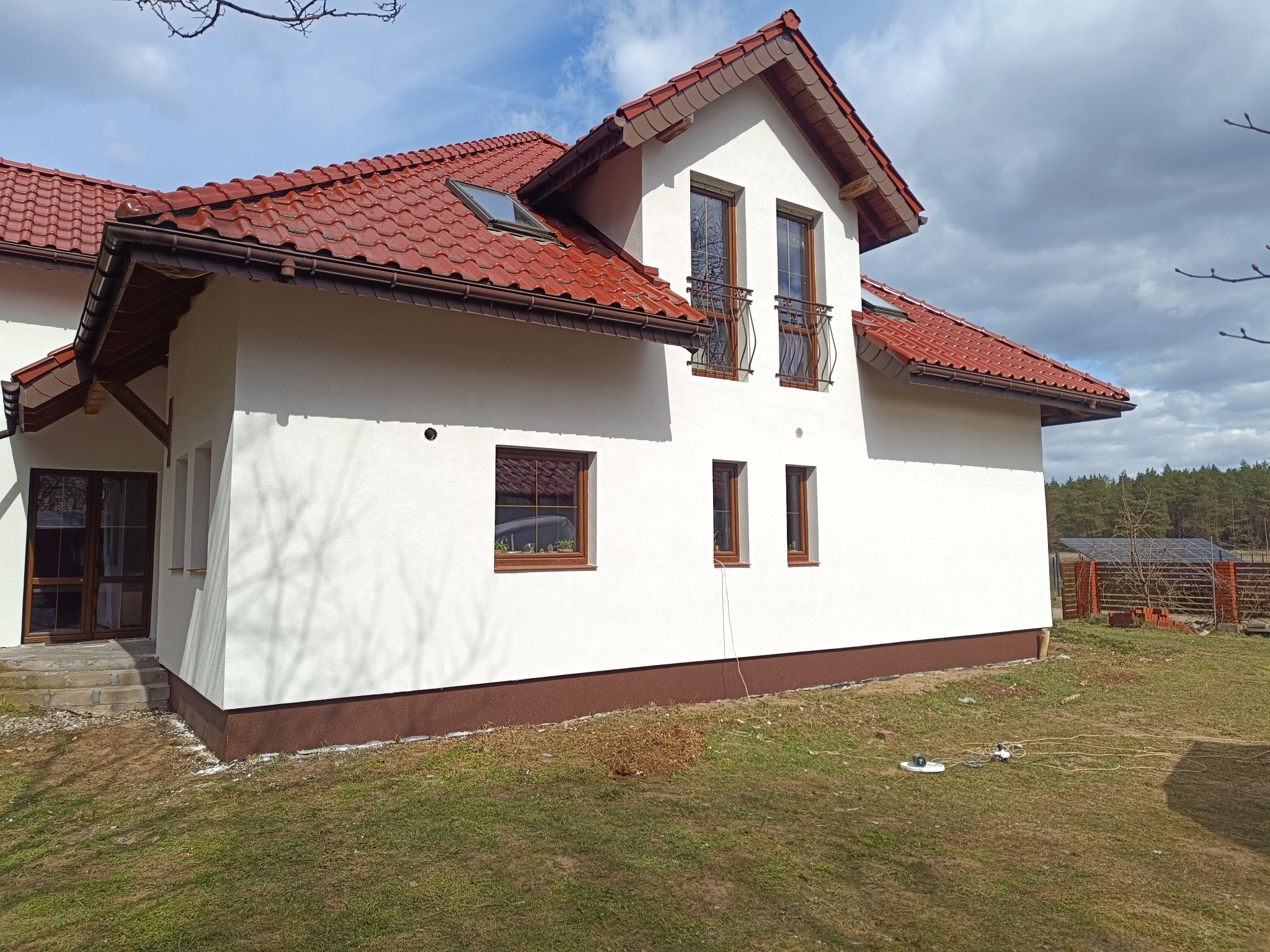 Biały dom z czerwoną dachówką, brązowymi oknami i wykończeniem. Widoczne detale architektoniczne i świeżo pomalowana elewacja. Tło stanowi błękitne niebo.