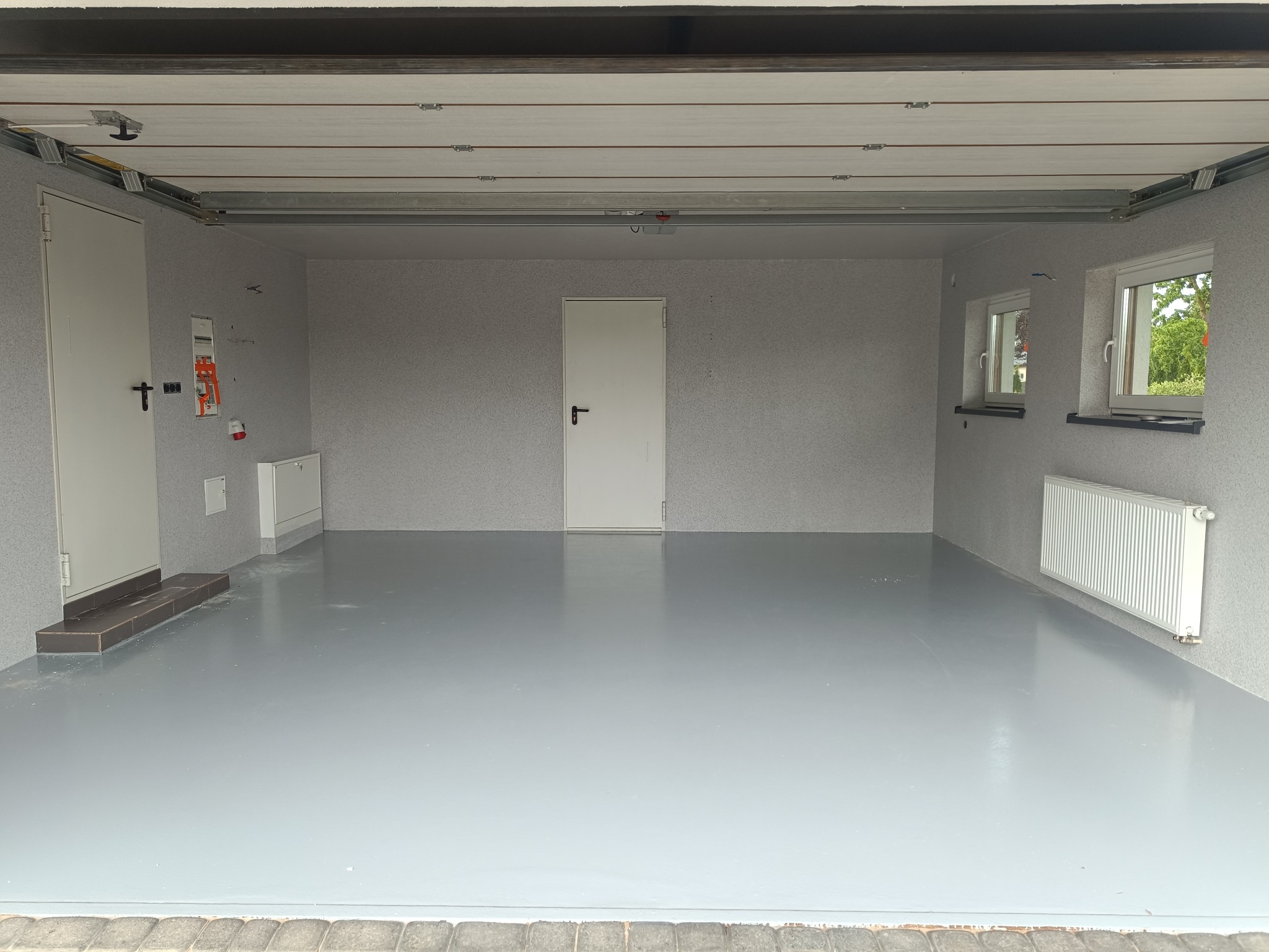 Wnętrze garażu po remoncie: szare ściany o fakturze baranka, gładka, szara posadzka żywiczna, białe drzwi i okna, grzejnik. Widok z zewnątrz, lekko z dołu.