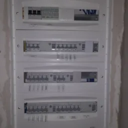 Rozdzielnica elektryczna z czterema rzędami zabezpieczeń, opisana etykietami wskazującymi obwody (pralka, rolety, zmywarka, oświetlenie górne, pompa ciepła), zamontowana na surowej ścianie...