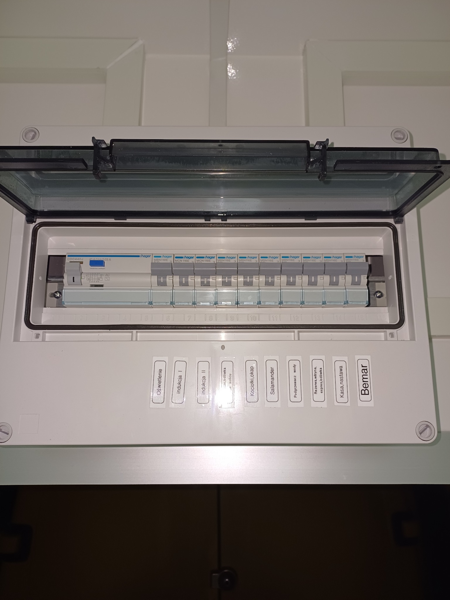 Otwarta skrzynka bezpiecznikowa Hager z opisami obwodów na naklejkach, widok z przodu, biała obudowa.