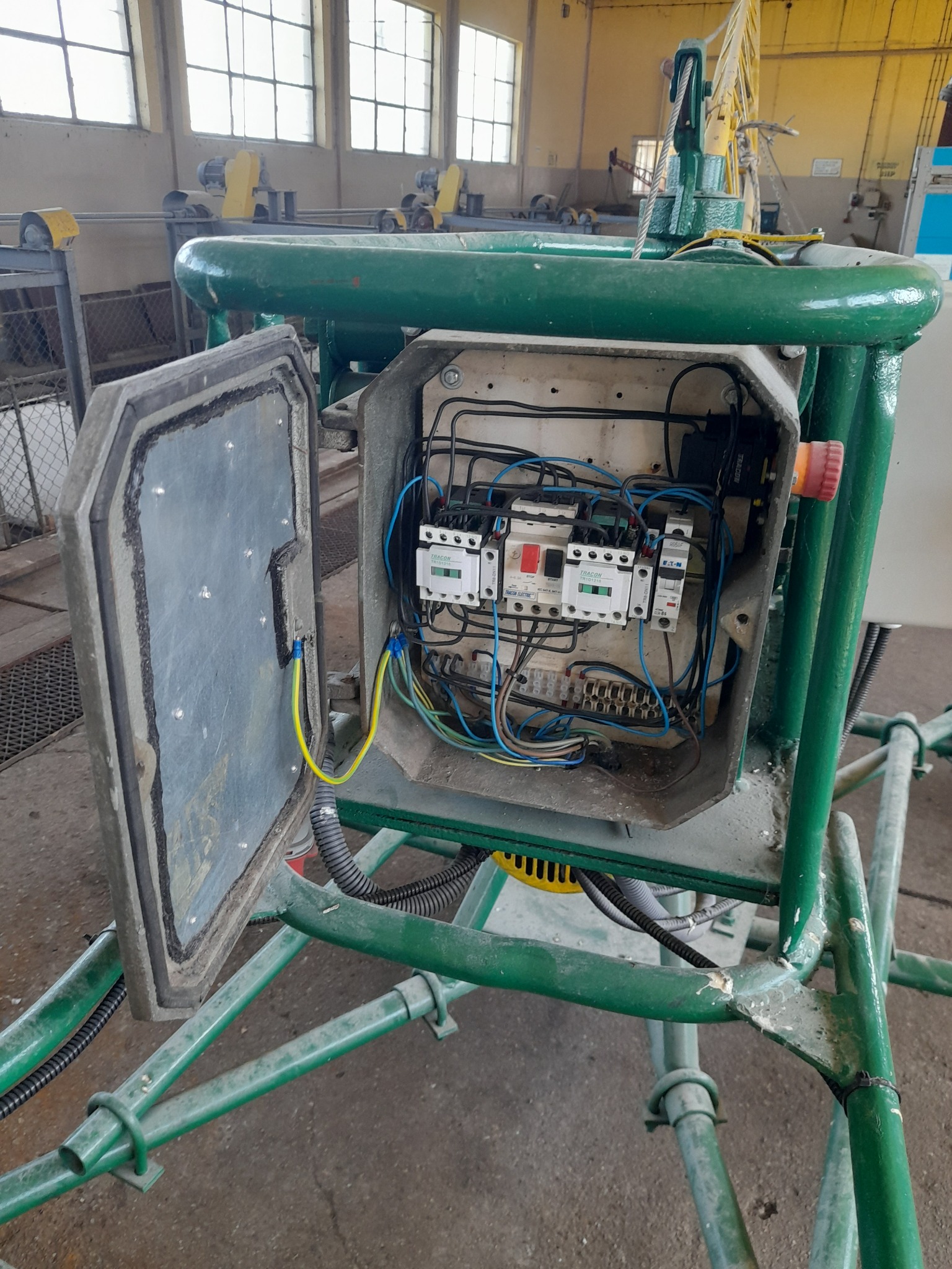 Otwarta skrzynka elektryczna z widocznymi bezpiecznikami, przewodami i elementami sterującymi, umieszczona na konstrukcji z zielonych rur w hali przemysłowej.