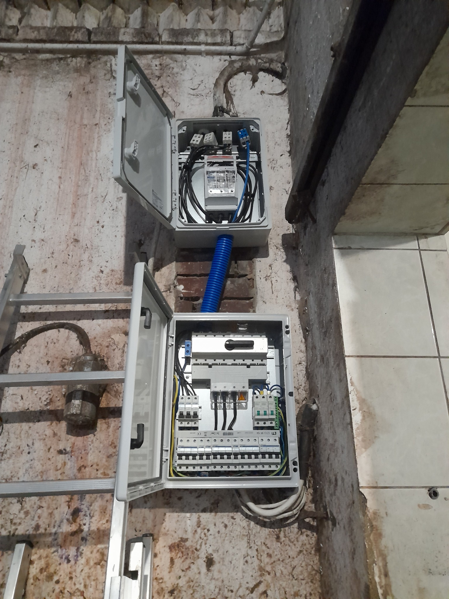 Dwa rozdzielnice elektryczne zamontowane na nierównej ścianie, po lewej drabina aluminiowa, widoczne kable i zabezpieczenia wewnątrz otwartych skrzynek, niebieska rura karbowana łączy obie...