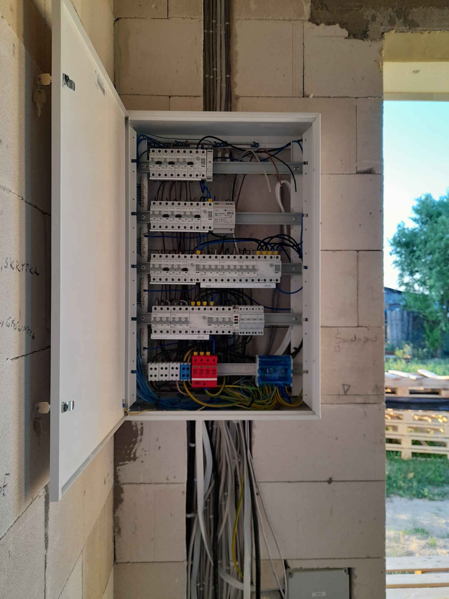 Otwarta skrzynka elektryczna z zamontowanymi bezpiecznikami i okablowaniem, zamontowana na ścianie z betonu komórkowego w surowym stanie.