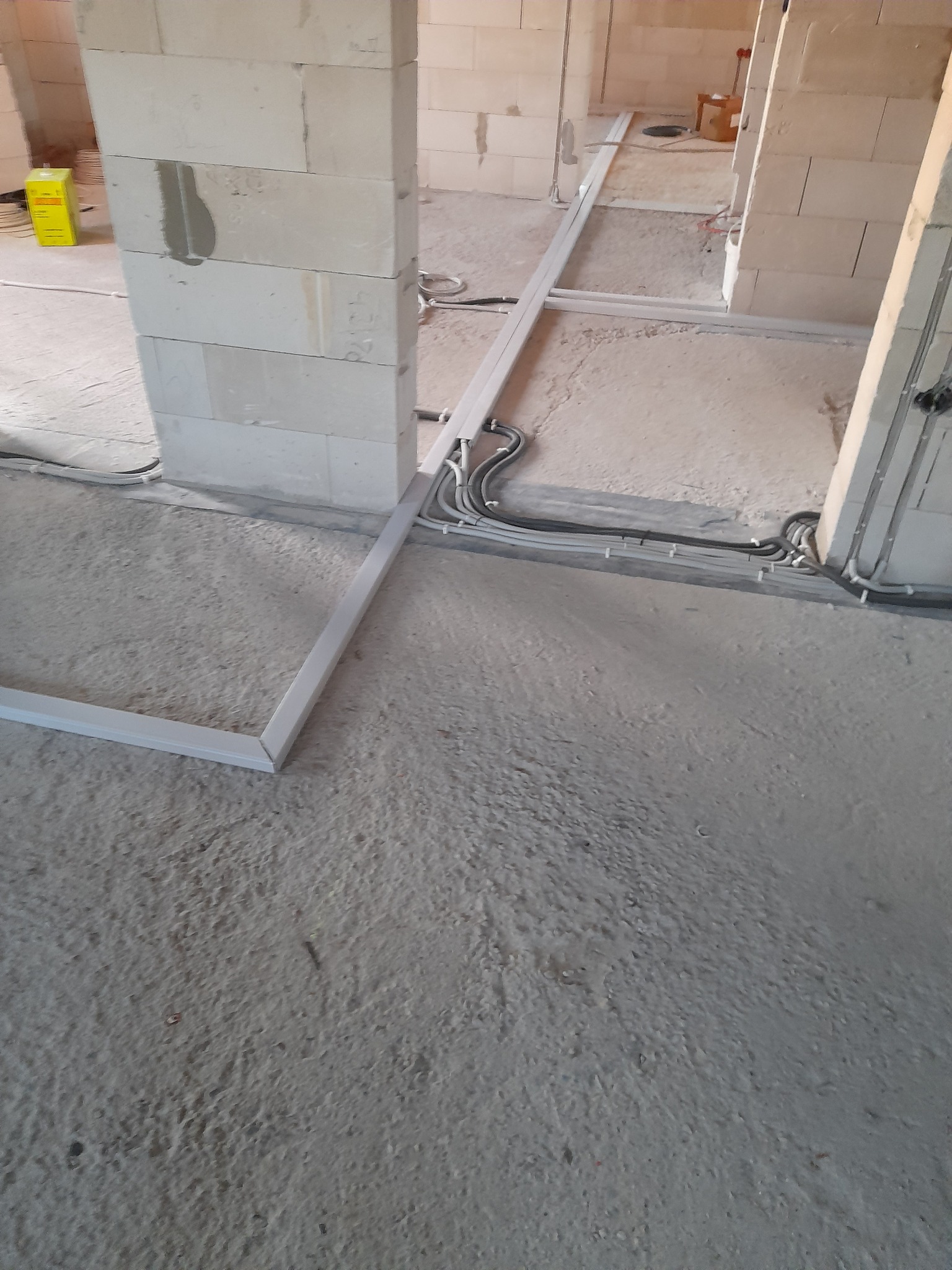 Surowy stan instalacji elektrycznej w budynku w trakcie budowy: widoczne kable i rury prowadzone po podłodze wzdłuż ścian z pustaków.