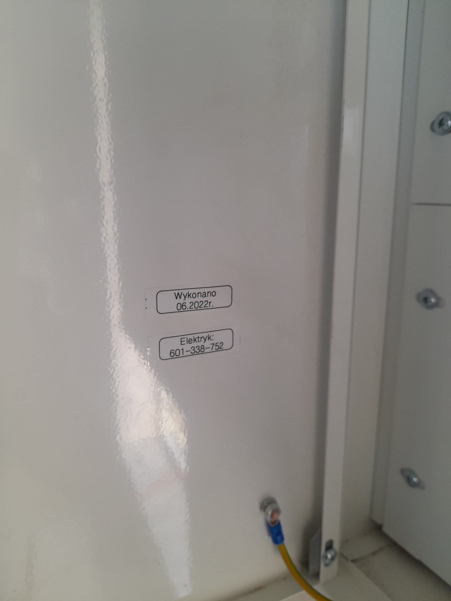 Wnętrze białej szafy elektrycznej z naklejkami informacyjnymi o wykonaniu prac (data 06.2022) i numerze telefonu do elektryka, widoczne przyłącze uziemiające z niebieską końcówką.