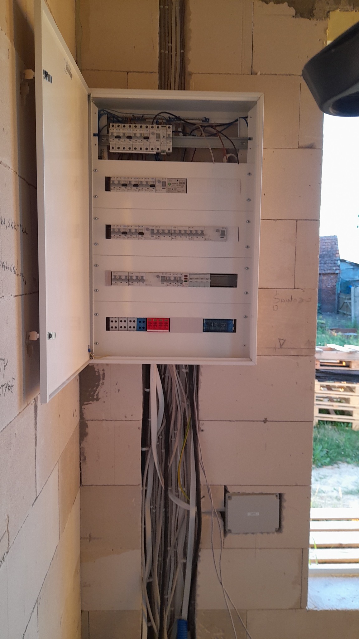 Otwarta skrzynka rozdzielcza z widocznymi bezpiecznikami i przewodami, zamontowana w surowej ścianie z betonu komórkowego, z widocznymi przewodami w bruździe pod skrzynką.