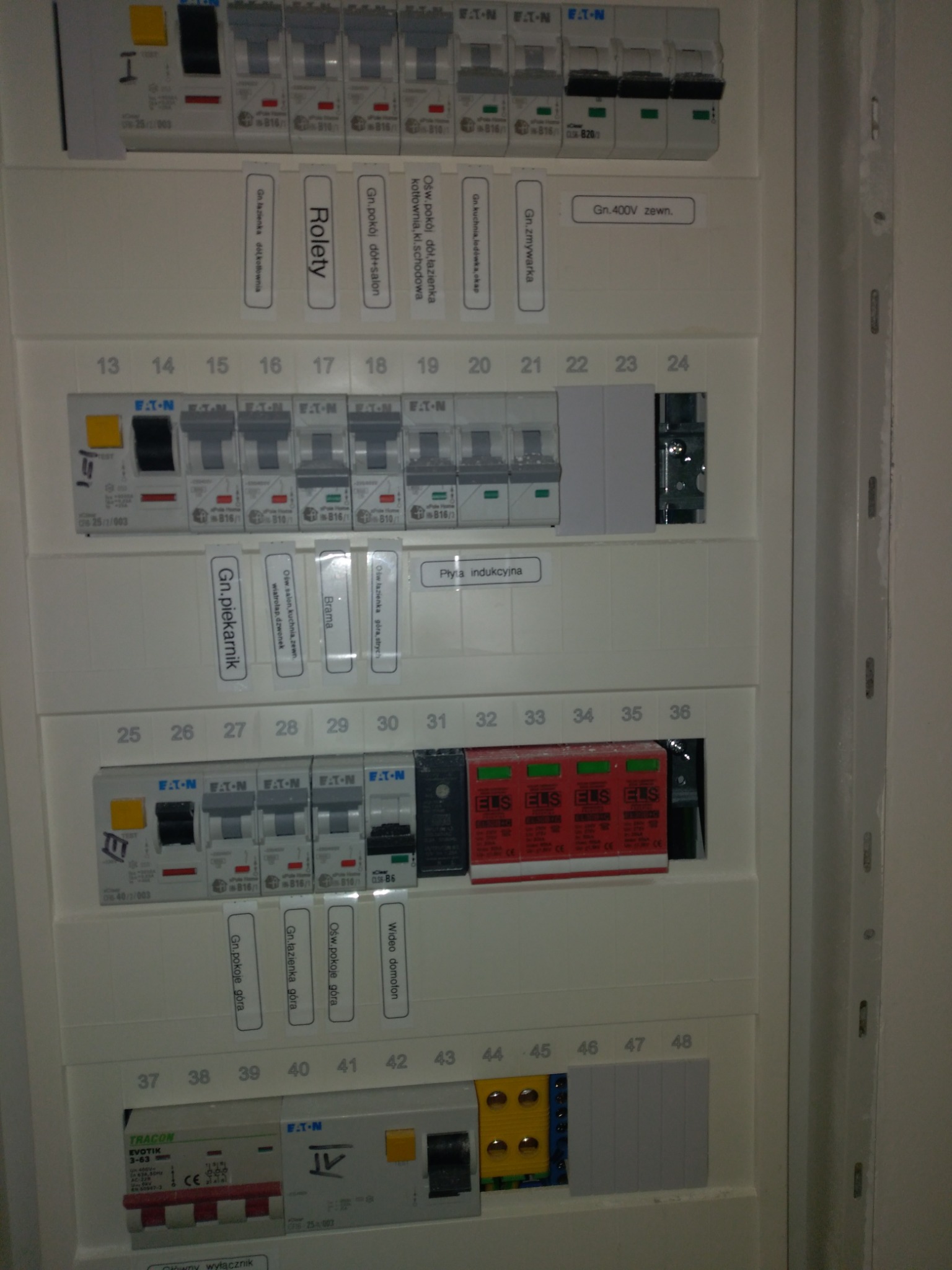Rozdzielnica elektryczna z licznymi bezpiecznikami i opisami obwodów, widoczne etykiety z nazwami pomieszczeń i urządzeń, takich jak 'Rolety', 'Płyta indukcyjna', 'Gn. piekarnik', 'Ośw. łazienka...