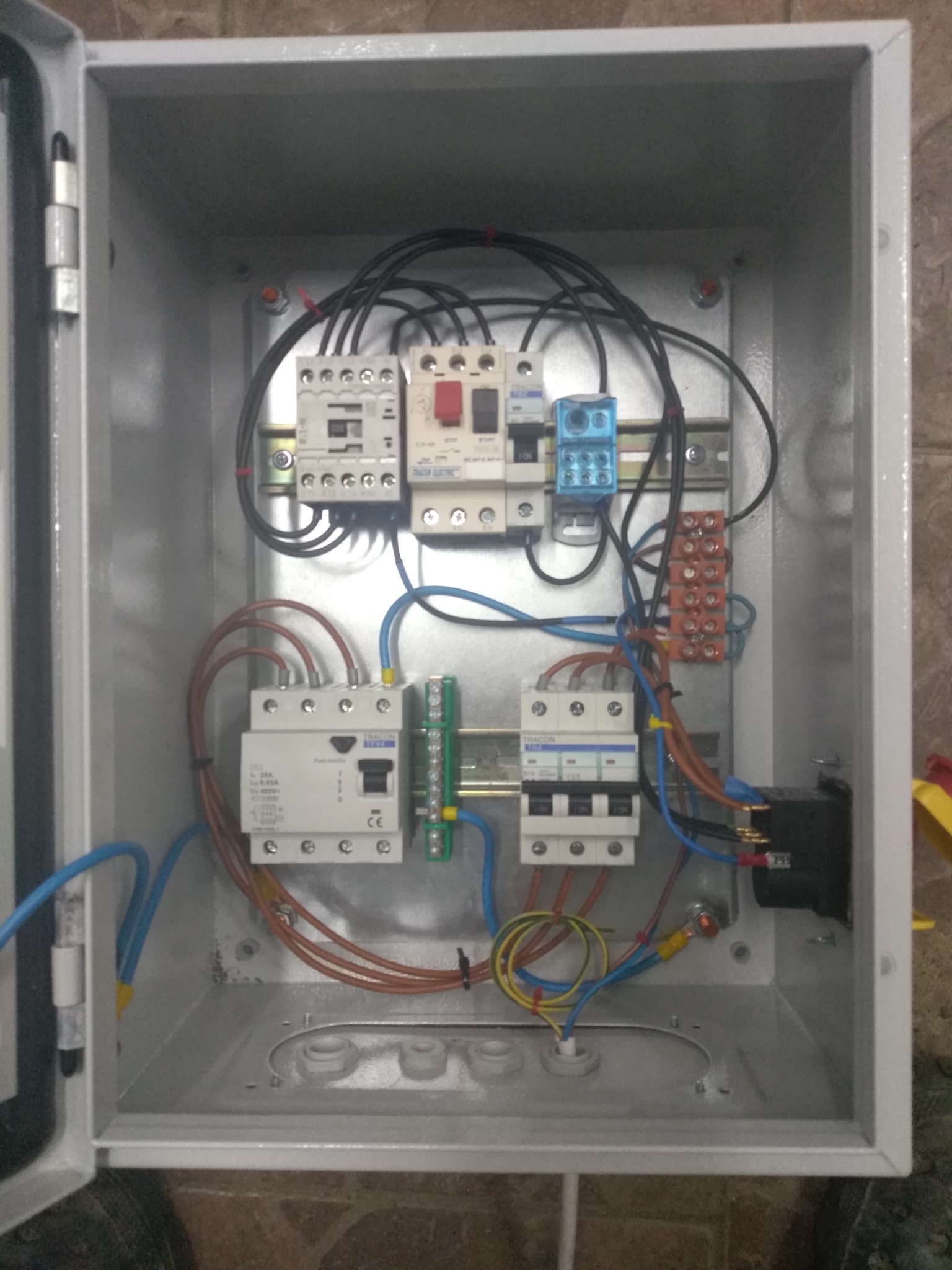 Otwarta skrzynka elektryczna z zamontowanymi bezpiecznikami, stycznikami i przewodami o różnych kolorach, zamocowanymi na szynach DIN.