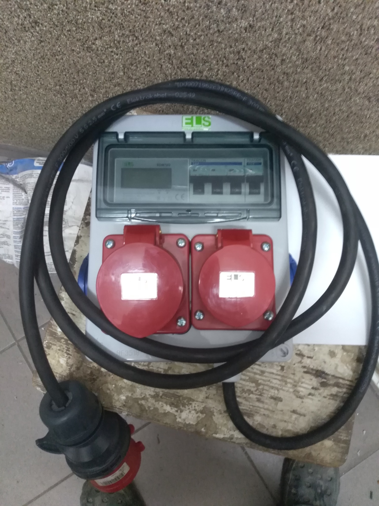 Rozdzielnica elektryczna z dwoma czerwonymi gniazdami przemysłowymi, zabezpieczona przezroczystą pokrywą, podłączona grubym, czarnym kablem zakończonym wtyczką. Widoczne oznaczenia na kablu...