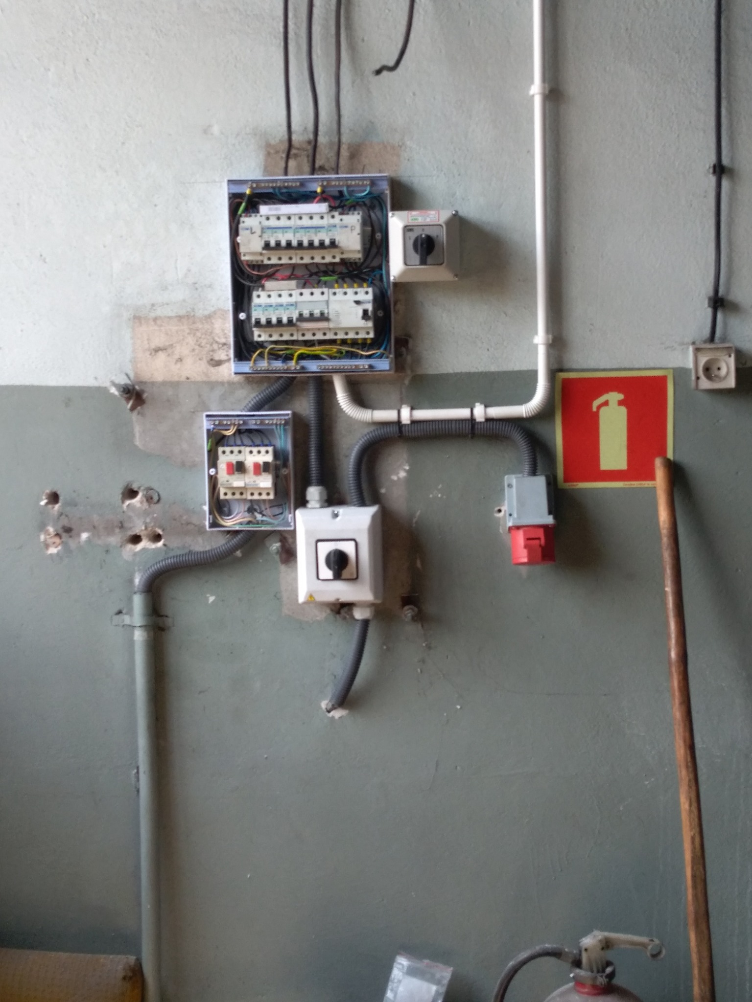Rozdzielnice elektryczne, wyłączniki i gniazdo siłowe zamontowane na szarej, częściowo odrapanej ścianie, widoczny znak przeciwpożarowy i gaśnica.