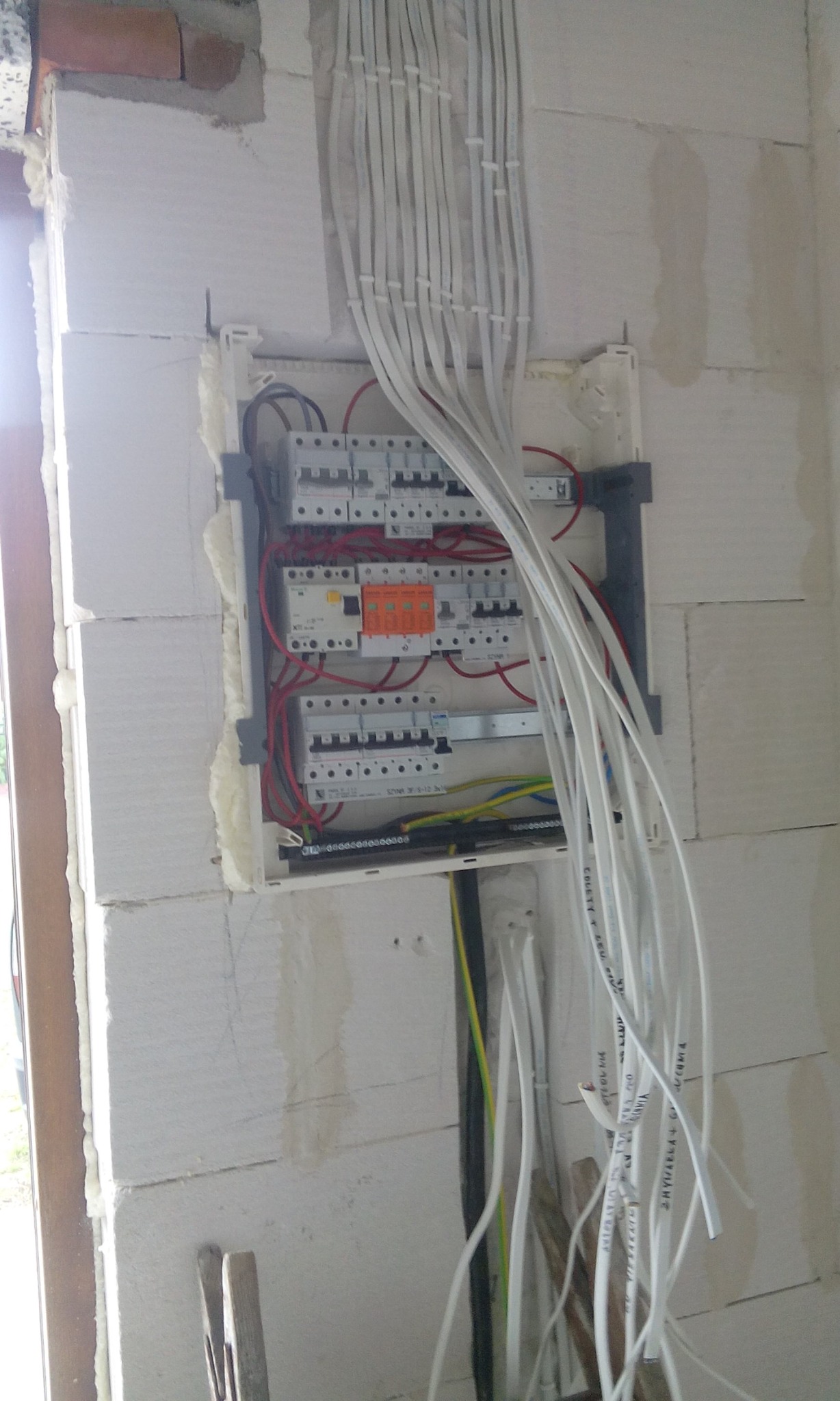 Rozdzielnica elektryczna z licznymi przewodami w surowym stanie, zamontowana na ścianie z białych bloczków, widoczne zabezpieczenia i przewody w różnych kolorach.