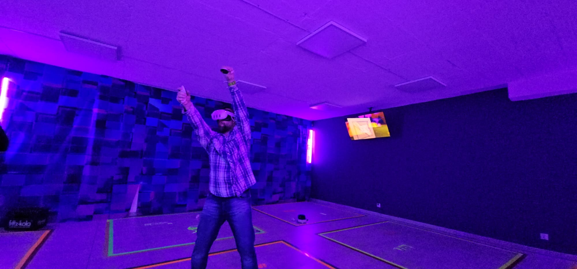 Mężczyzna w okularach VR i kontrolerach w rękach, stojący w fioletowym pomieszczeniu z neonami i ekranem telewizora, prawdopodobnie podczas gry.