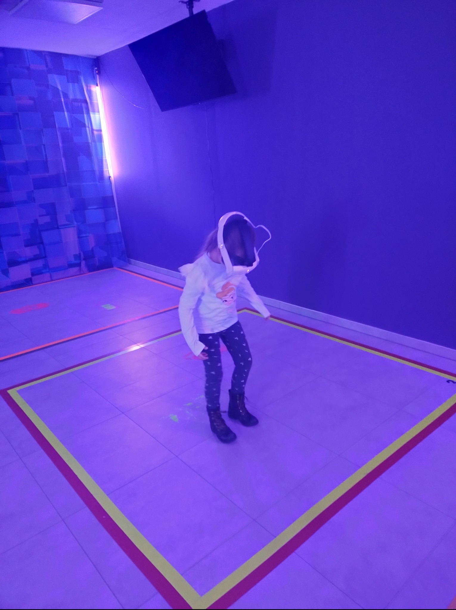 Dziewczynka w okularach VR w pokoju z podświetlonymi kwadratami na podłodze i ścianą z dekoracyjnymi panelami 3D, oświetlonymi fioletowym światłem.