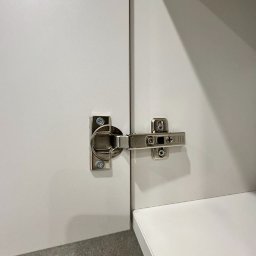 Eryk Kurek - Szczegółowe ujęcie zawiasu metalowego zamontowanego na białych drzwiczkach szafki, widoczne wkręty mocujące.