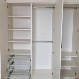 Biała szafa wnękowa z otwartymi drzwiami, widoczne półki, szuflady koszykowe i drążki na ubrania. Jasne wnętrze, drewniana podłoga.