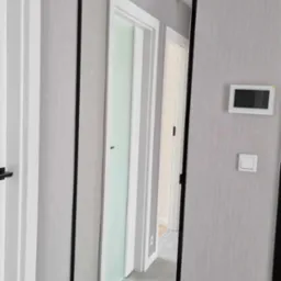 Wysokie lustro w czarnej ramie, zamontowane w przedpokoju z szarą tapetą i białymi drzwiami. Widoczne odbicie wnętrza mieszkania.