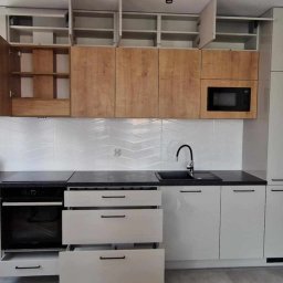 Eryk Kurek - Nowoczesna kuchnia z otwartymi szafkami górnymi i drewnianymi frontami, czarny blat, białe płytki chevron i otwartą szufladą, minimalistyczny design.