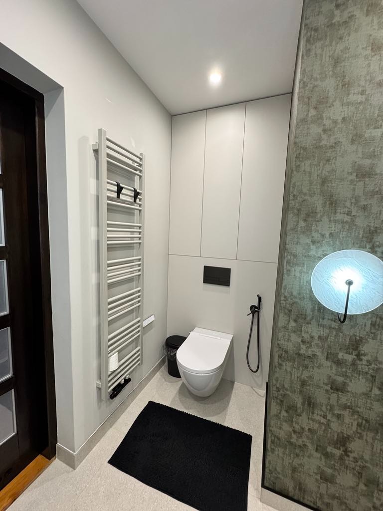 Nowoczesna łazienka z zabudowaną szafą w neutralnych kolorach, wisząca toaleta z bidetką, grzejnik drabinkowy na ścianie i czarny dywanik.