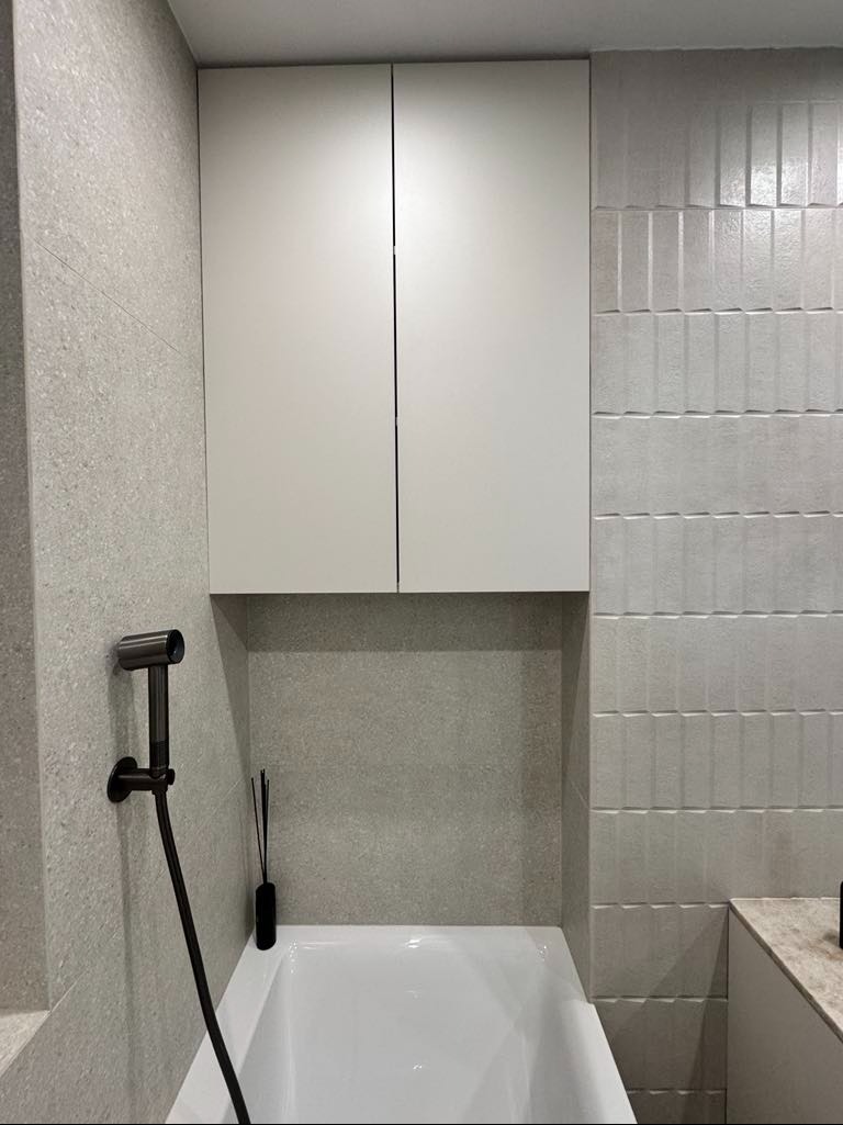 Biała szafka wisząca nad wanną, dopasowana kolorystycznie do szarej ściany z płytek, z widocznym czarnym bidetem i dyfuzorem zapachowym.