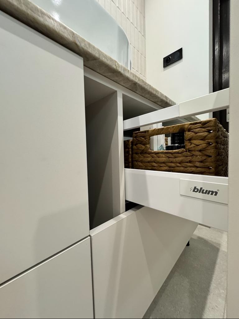 Biała szafka łazienkowa z wysuwaną szufladą wyposażoną w koszyk z wikliny, widoczne okucia Blum.