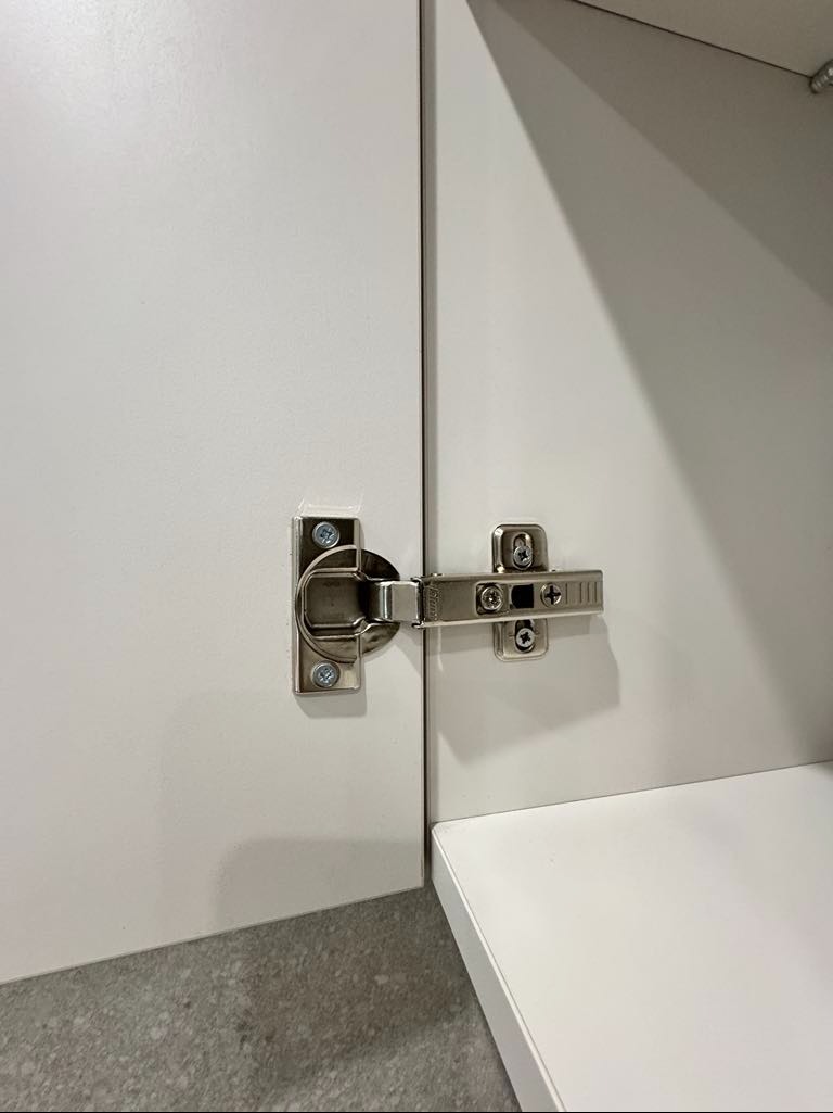 Szczegółowe ujęcie zawiasu metalowego zamontowanego na białych drzwiczkach szafki, widoczne wkręty mocujące.