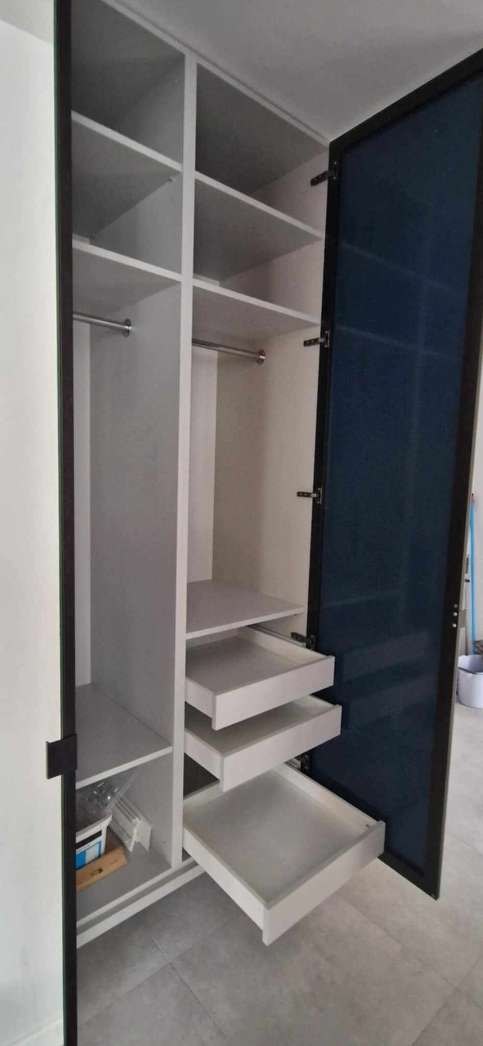 Wnętrze szafy wnękowej z otwartymi drzwiami i wysuniętymi szufladami. Białe półki i szuflady kontrastują z ciemną ramą drzwi.