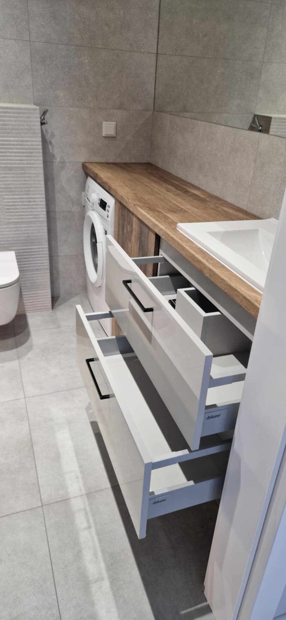 Nowoczesna łazienka z drewnianym blatem, białą umywalką i szafkami z szufladami. Obok pralka i toaleta. Szare płytki na ścianach i podłodze.