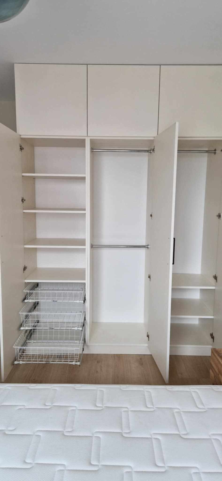 Biała szafa wnękowa z otwartymi drzwiami, widoczne półki, szuflady koszykowe i drążki na ubrania. Jasne wnętrze, drewniana podłoga.