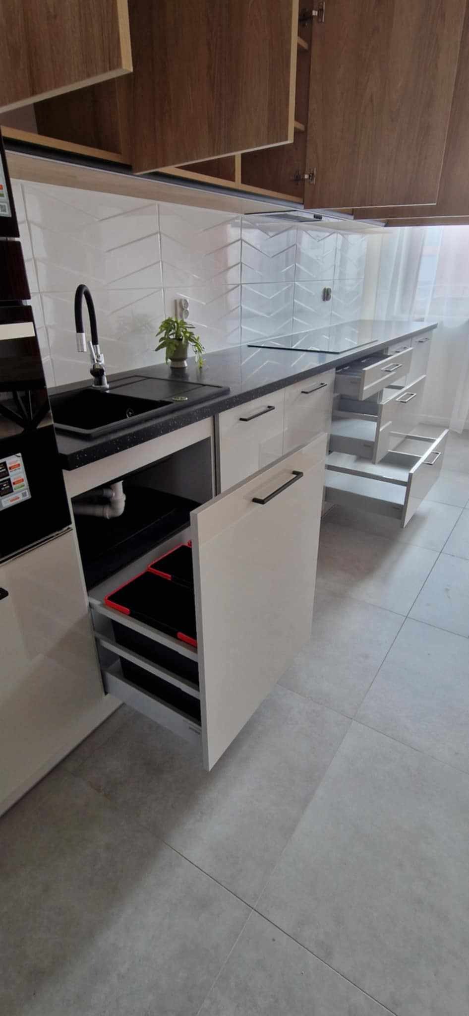 Biała zabudowa kuchenna z czarnym blatem i wysuwanymi szufladami, otwarty front szafki z koszami na śmieci, nowoczesny design, płytki w jodełkę na ścianie.