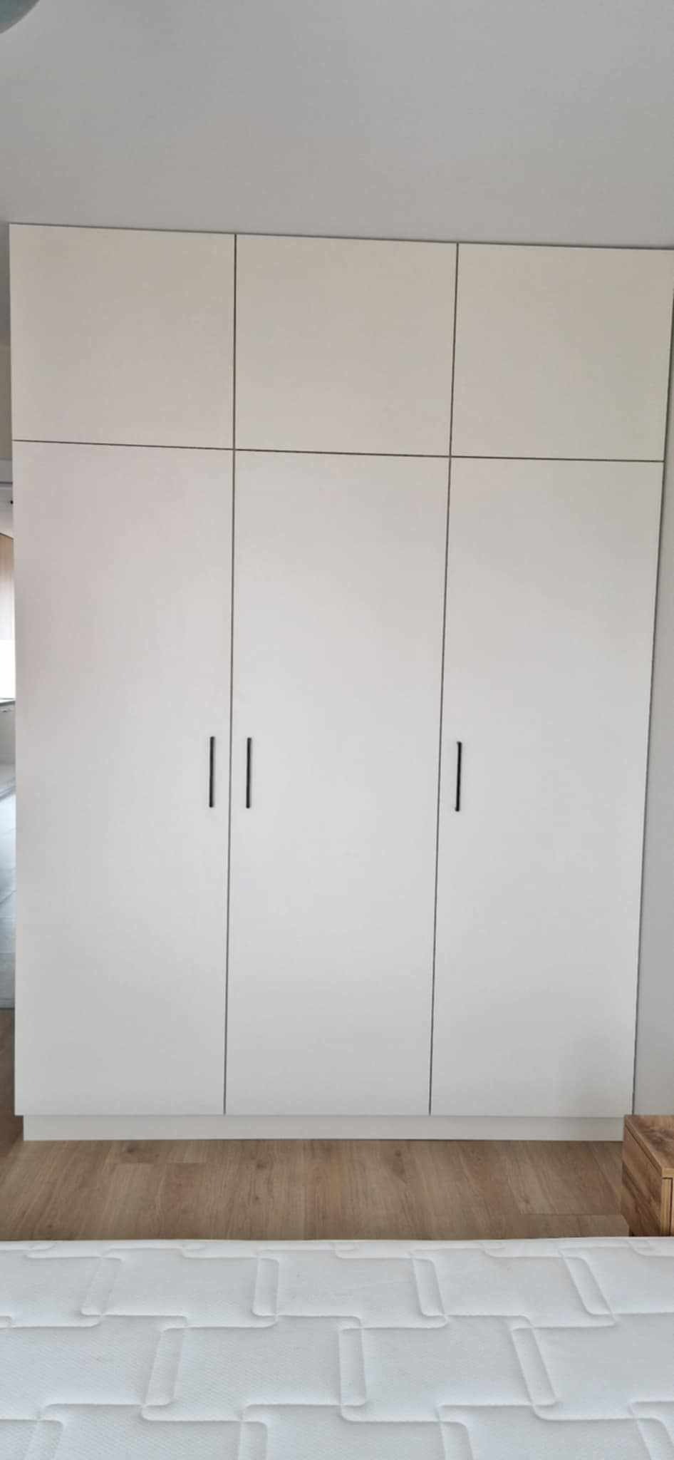 Biała szafa wnękowa z czarnymi, minimalistycznymi uchwytami, dopasowana do jasnego wnętrza z drewnianą podłogą. Prosty design i funkcjonalność.