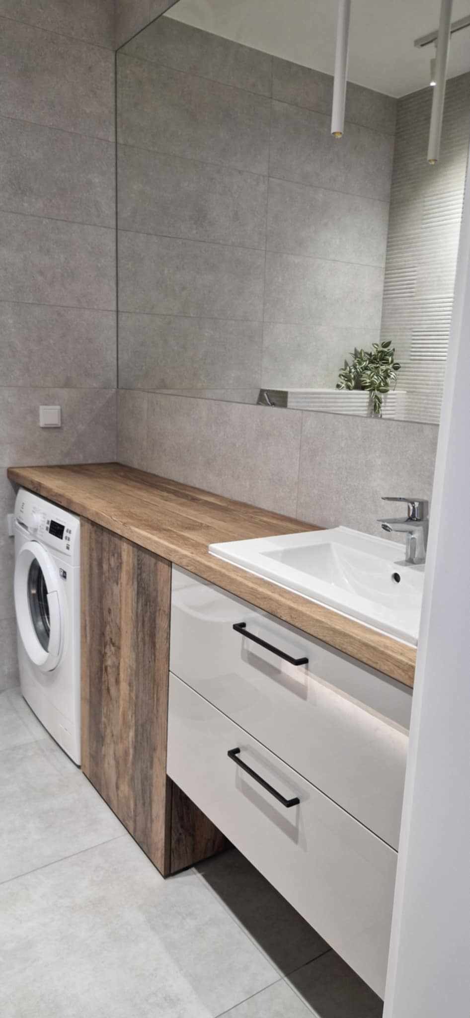 Nowoczesna łazienka z zabudową pralki i umywalki z szufladami, drewniany blat, szare płytki. Minimalistyczny design, oświetlenie LED pod szafkami.