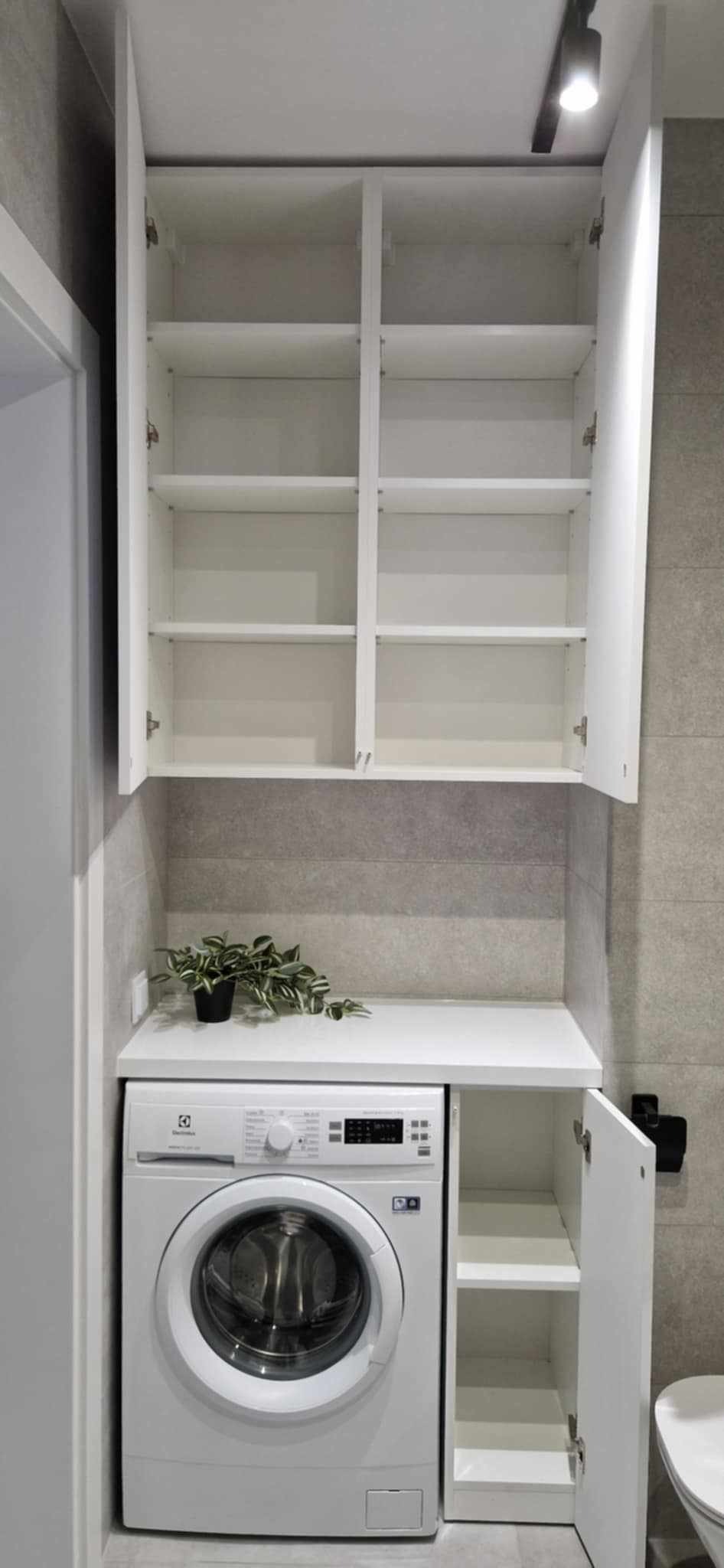 Biała pralka Electrolux z szafką nad nią w łazience z szarymi płytkami. Szafka ma otwarte drzwiczki, a na blacie stoi doniczka z rośliną.