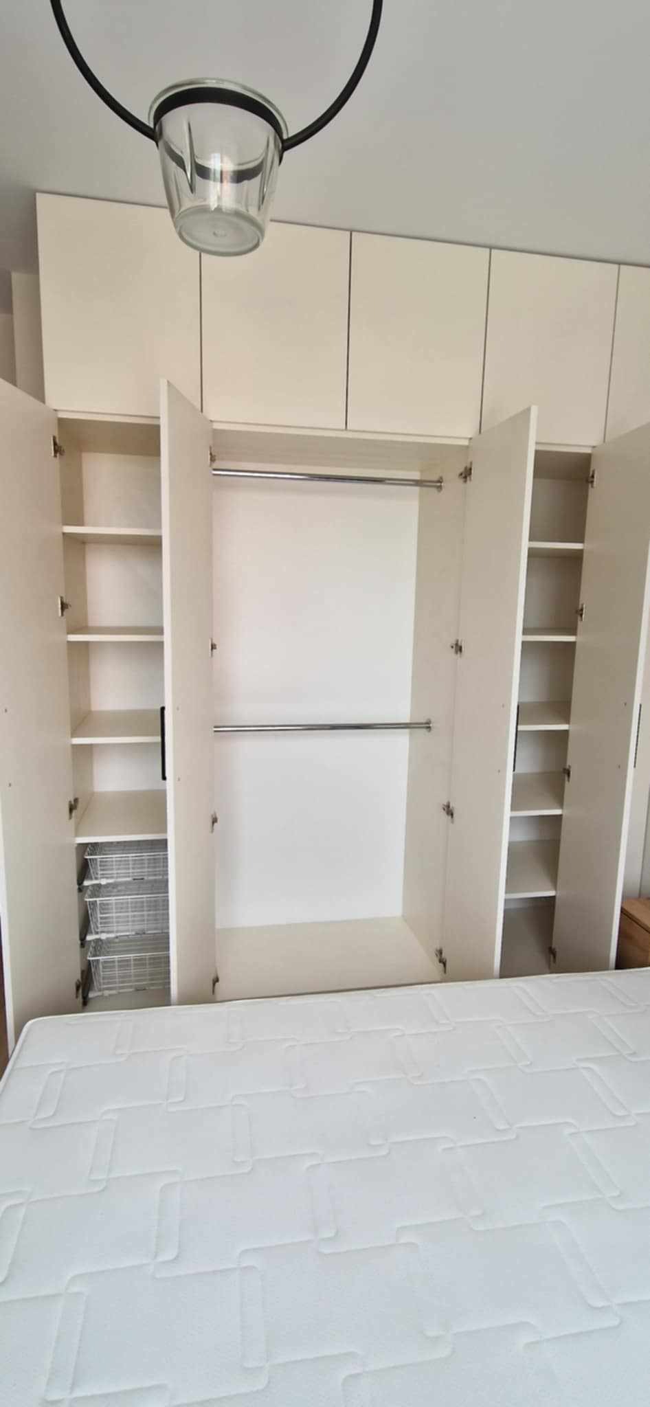 Biała szafa wnękowa z otwartymi drzwiami, widoczne półki i drążki na ubrania. Na dole materac. Górne szafki zamknięte, pod sufitem lampa.