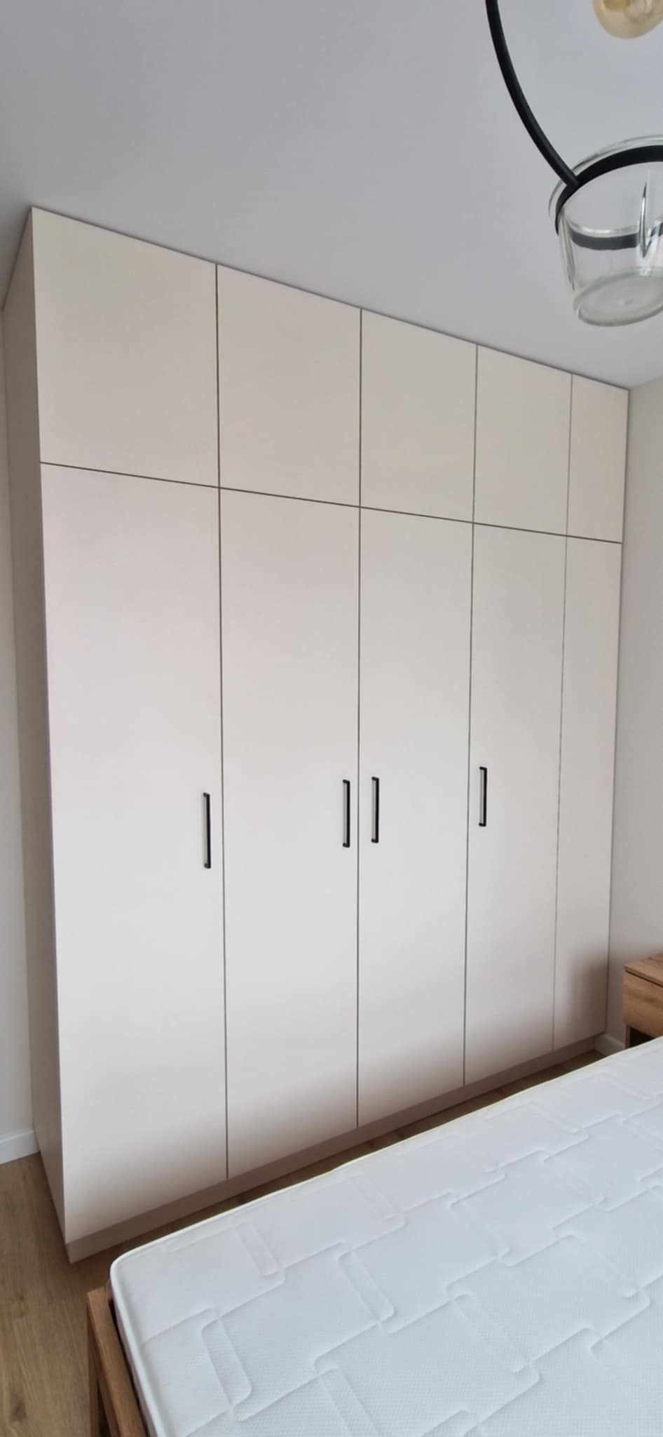 Jasna szafa wnękowa z czarnymi, pionowymi uchwytami, zabudowana od podłogi do sufitu w sypialni, widok z perspektywy łóżka.