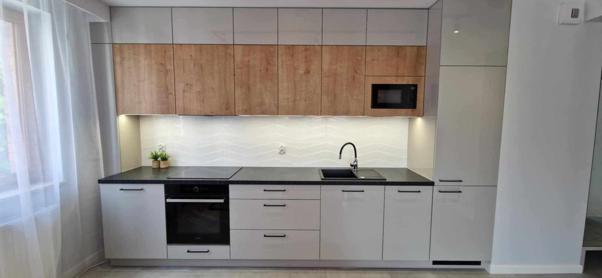 Nowoczesna kuchnia z białymi szafkami dolnymi i drewnopodobnymi górnymi, czarnym blatem i sprzętem AGD. Minimalistyczny design z subtelnym oświetleniem podszafkowym.