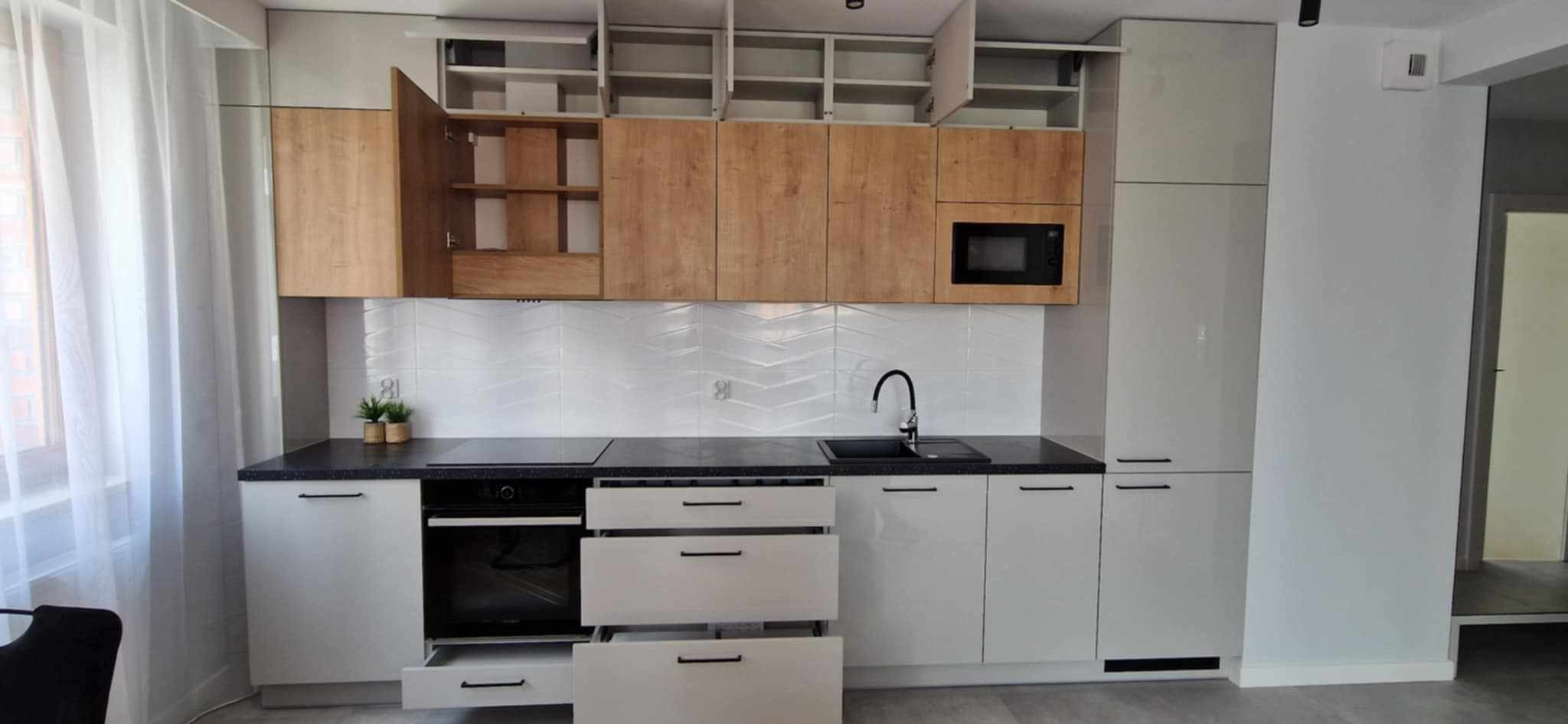 Nowoczesna kuchnia z otwartymi szafkami górnymi i drewnianymi frontami, czarny blat, białe płytki chevron i otwartą szufladą, minimalistyczny design.