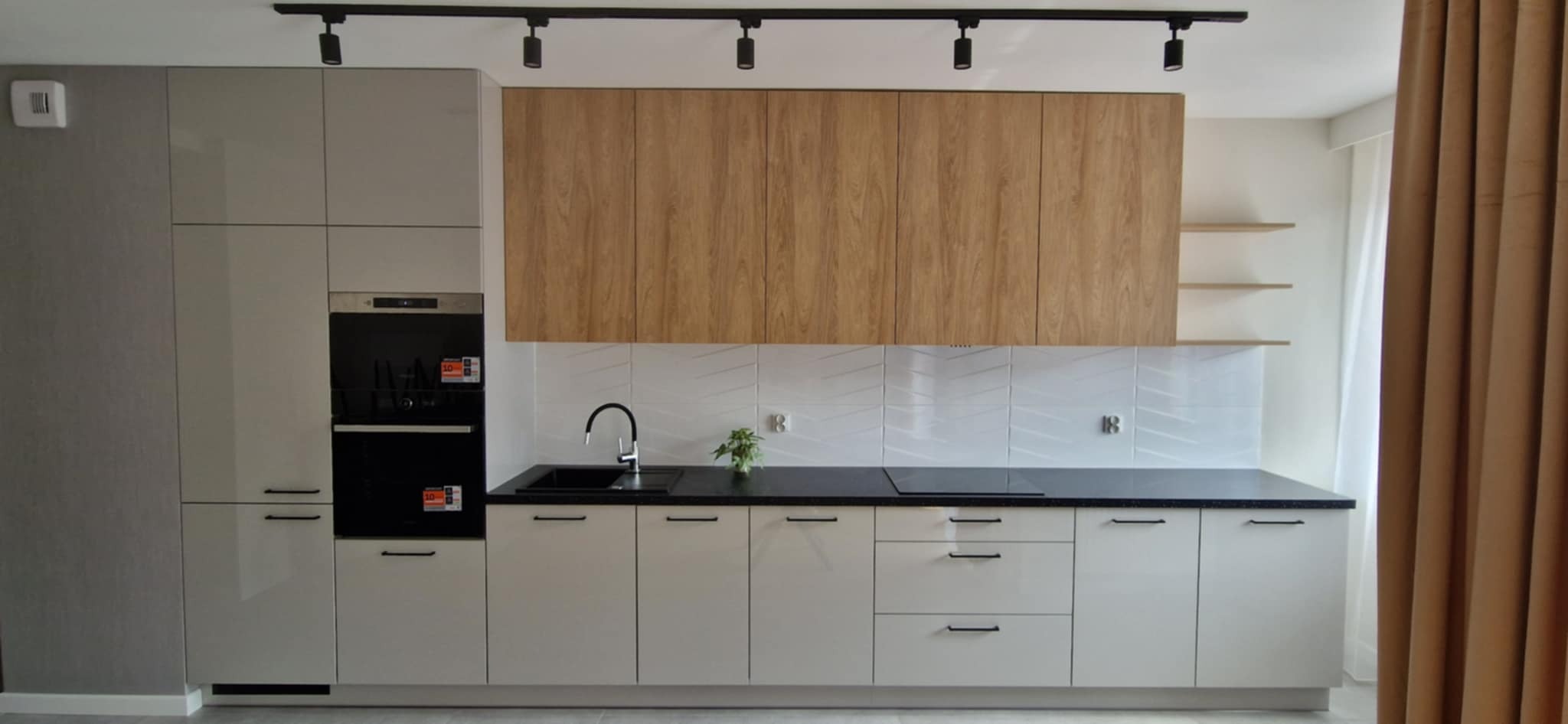 Nowoczesna kuchnia na wymiar z drewnianymi górnymi szafkami, czarnym blatem i białymi, geometrycznymi płytkami na ścianie. Oświetlenie szynowe.