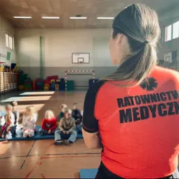 Instruktorka w czerwonej koszulce z napisem 'Ratownictwo Medyczne' prowadzi zajęcia z pierwszej pomocy dla grupy dzieci siedzących na podłodze sali gimnastycznej.