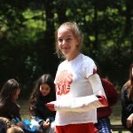 Uśmiechnięta dziewczyna z zabandażowaną ręką prezentuje prawidłowo założony opatrunek trójkątny na tle grupy osób i zalesionego terenu.