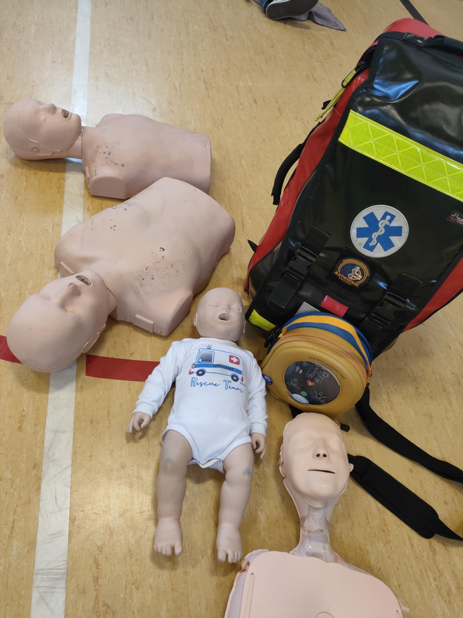 Fantomy do nauki resuscytacji, w tym manekin niemowlęcia w body z napisem 'Rescue Team', obok torby ratowniczej z symbolem Eskulapa i defibrylatora.