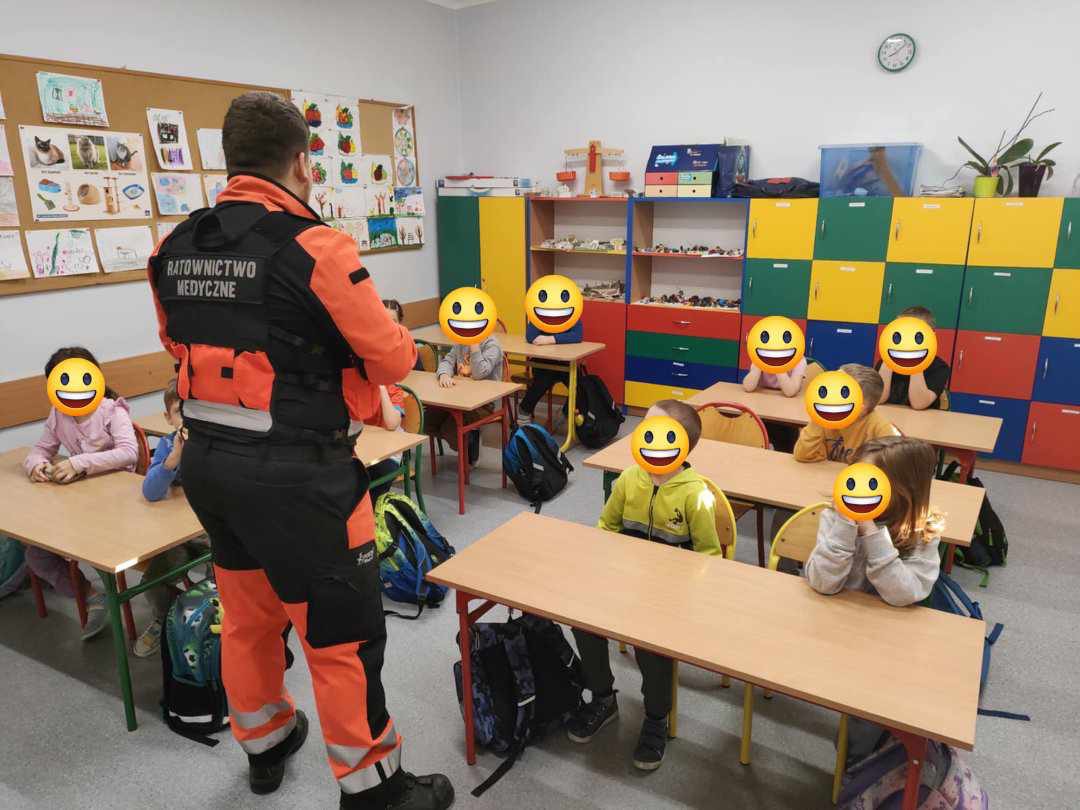 Instruktor ratownictwa medycznego, ubrany w pomarańczowo-czarny strój z napisem 'RATOWNICTWO MEDYCZNE' na plecach, prowadzi zajęcia dla dzieci w klasie szkolnej. Dzieci siedzą przy biurkach.
