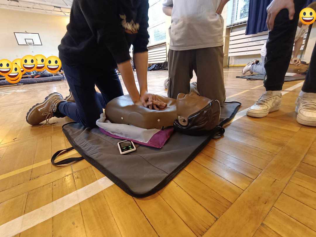 Osoba w trakcie ćwiczeń resuscytacji krążeniowo-oddechowej na manekinie na sali gimnastycznej, widoczny AED na macie, inni uczestnicy w tle.