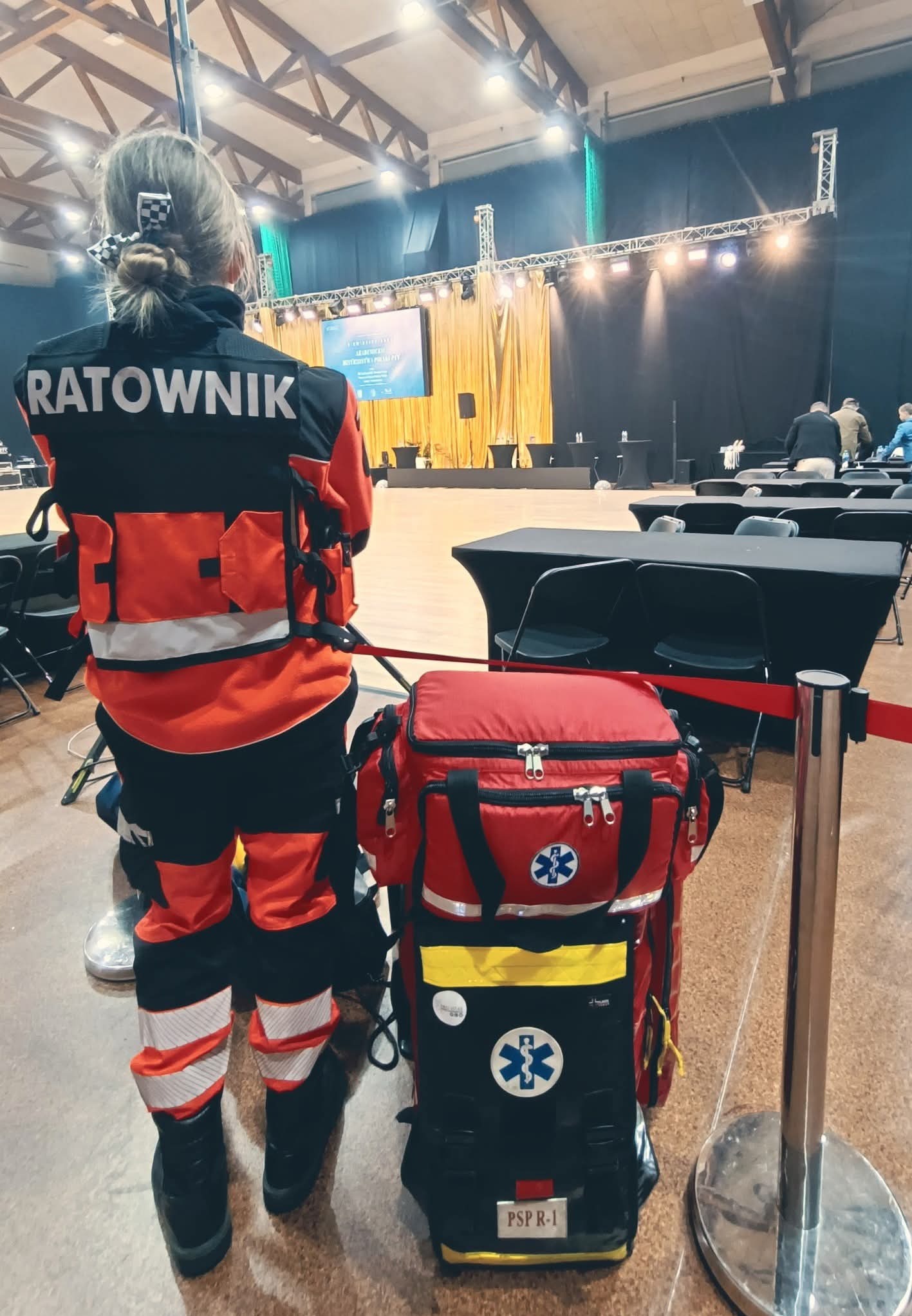 Kobieta ratownik w stroju z torbą medyczną PSP R-1, tyłem do widza, na tle sali konferencyjnej z ustawionymi stołami. Szkolenie z pierwszej pomocy.