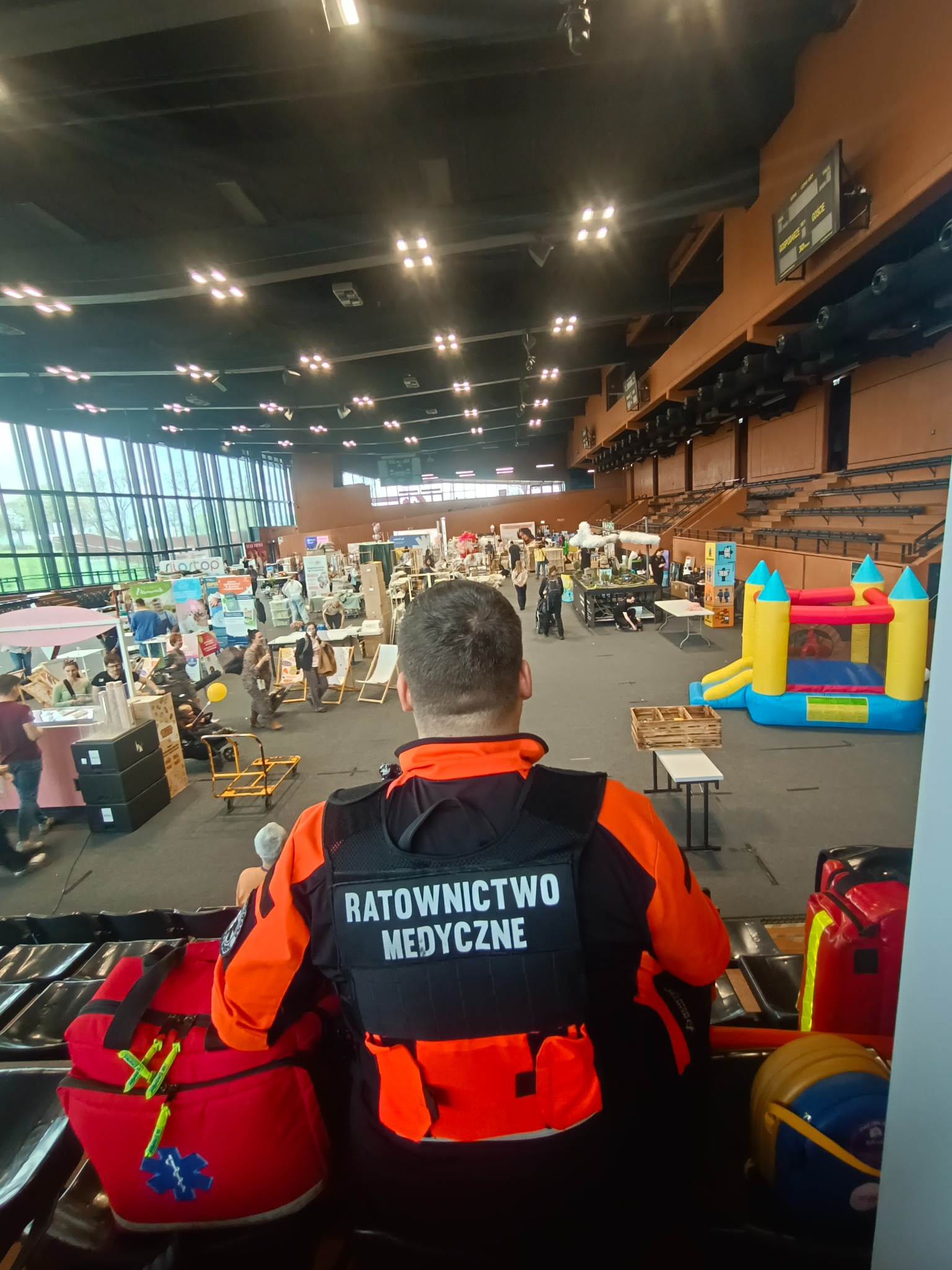 Ratownik medyczny w pomarańczowej kurtce, z widokiem na halę targową z dmuchanym zamkiem. Torba medyczna na pierwszym planie.